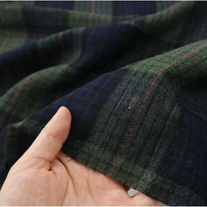 May include: Close-up of a folded fabric with a plaid pattern. The fabric features a combination of dark green, navy blue, and red lines. The fabric is held by a hand, showing the texture and drape of the material.