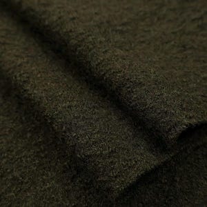 May include: A dark olive green fabric with a thick, fuzzy, and textured surface. The material is folded over itself, creating a layered appearance that highlights the dense, fibrous quality of the textile and its deep, uniform colour throughout.
