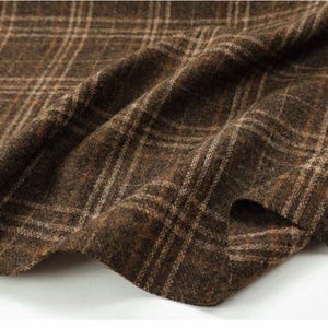 Puede incluir: Una tela de color marrón oscuro con una superficie tejida y texturizada que presenta un sutil patrón de cuadros con líneas en tonos beige y crema. El material está drapeado en pliegues suaves, resaltando su consistencia gruesa y el diseño.