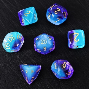 Könnte beinhalten: Ein Set aus sieben polyedrischen Würfeln mit einem durchscheinenden Farbverlauf in Blau und Violett. Jeder Würfel hat scharfe, facettierte Kanten und ist mit goldfarbenen Zahlen beschriftet. Das Set enthält verschiedene Formen, darunter ein zwanzigseitiger Würfel.