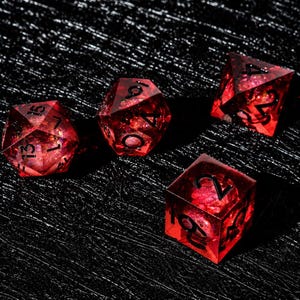 Peut inclure: Trois d&eacute;s poly&eacute;driques rouge translucide avec des paillettes incrust&eacute;es et des chiffres &agrave; l'encre noire. L'ensemble comprend un d&eacute; &agrave; vingt faces, un d&eacute; &agrave; douze faces et un d&eacute; cubique &agrave; six faces, tous dot&eacute;s d'ar&ecirc;tes vives et d'une finition brillante.