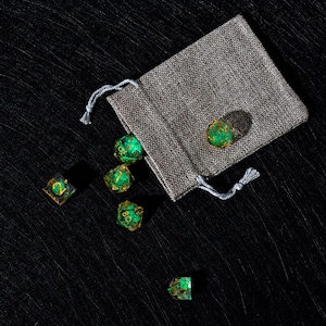 Peut inclure: Un ensemble de d&eacute;s poly&eacute;driques vert translucide avec des chiffres dor&eacute;s, contenant des inclusions scintillantes &agrave; l'int&eacute;rieur de chaque d&eacute;. Les d&eacute;s sont repr&eacute;sent&eacute;s sortant d'une petite pochette rectangulaire en tissu gris &agrave; cordon.