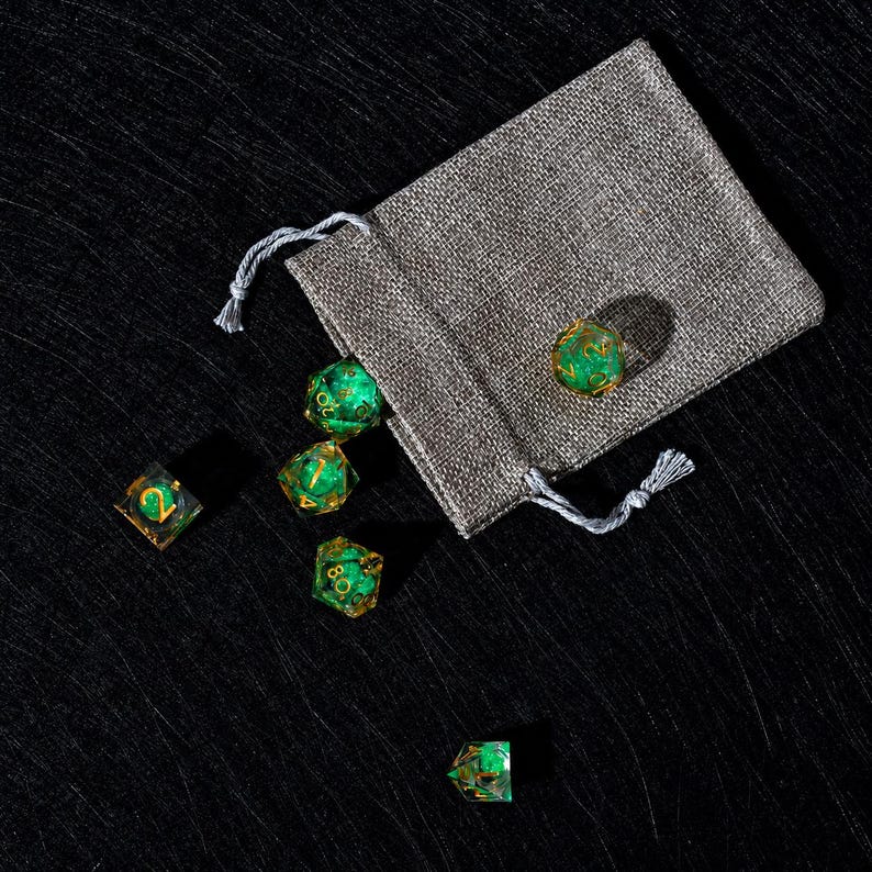 Peut inclure: Un ensemble de d&eacute;s poly&eacute;driques vert translucide avec des chiffres dor&eacute;s, contenant des inclusions scintillantes &agrave; l'int&eacute;rieur de chaque d&eacute;. Les d&eacute;s sont repr&eacute;sent&eacute;s sortant d'une petite pochette rectangulaire en tissu gris &agrave; cordon.