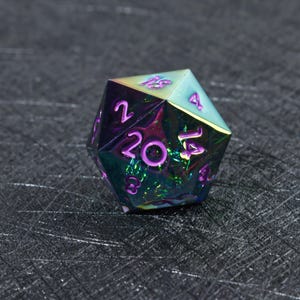May include: A twenty-sided die featuring an iridescent, multi-colored metallic finish that shifts between shades of purple, green, and blue. The numbers are engraved into the surface and filled with a bright purple pigment for high visibility.