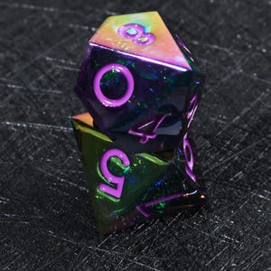 May include: Two stacked polyhedral dice with a metallic, iridescent finish that shifts between purple, green, and gold. The dice feature deep purple, recessed numerals including an eight, a zero, a four, a five, and a seven clearly visible.