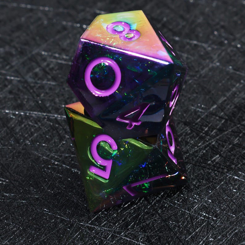 May include: Two stacked polyhedral dice with a metallic, iridescent finish that shifts between purple, green, and gold. The dice feature deep purple, recessed numerals including an eight, a zero, a four, a five, and a seven clearly visible.