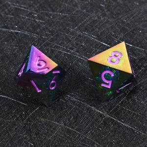 May include: Two multi-faceted polyhedral dice featuring an iridescent, rainbow-colored metallic finish. The dice have sharp edges and deep purple engraved numbers, including a visible 6, 9, 5, and 8, with a dark, reflective, and textured surface.