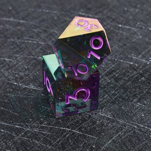 May include: A multi-faceted polyhedral die with an iridescent, oil-slick finish and deep purple engraved numbers. The die features a sharp-edged geometric design with a reflective surface that shifts between green, blue, and purple hues in the light.