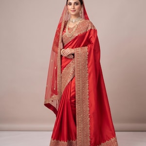 May include: A vibrant red satin saree featuring intricate gold embroidery along the borders. The ensemble includes a matching red sheer veil with gold detailing, a coordinating blouse with embroidered sleeves, and layered gold jewelry.