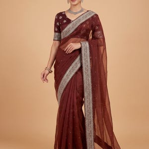 May include: A maroon saree with a matching blouse. The saree features a silver embroidered border and scattered embellishments. The blouse has short sleeves and a V-neck. The fabric appears sheer and lightweight, suitable for formal occasions.