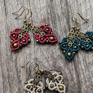 Peut inclure: Trois paires de boucles d'oreilles en macramé avec des motifs de cordons noués complexes en rouge foncé, bleu sarcelle et crème. Chaque paire comprend des fleurs en métal couleur laiton vieilli et de petites perles métalliques sur des crochets.