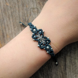 May include: A dark teal macramé bracelet featuring a central silver-toned metal floral charm and small silver-toned beads woven into the cord. The bracelet has an adjustable sliding knot closure with two dangling cord ends finished with silver beads.