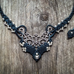 Op de afbeelding: Een donkerblauwe macramé ketting met een centraal zilverkleurig filigraan metalen onderdeel. Het koord is ingewikkeld geknoopt en versierd met talloze kleine, ronde, metallic zilveren kralen die langs de zijkanten en aan de onderkant zijn geplaatst.