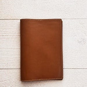 May include: A rectangular brown leather notebook cover featuring smooth, matte-textured leather with visible light-coloured stitching along all four edges. The cover is closed, displaying a uniform, solid brown colour across its entire front surface.