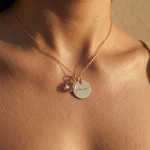 May include: A gold-colored necklace with a delicate chain. It features a small, round pendant with the name "jasmin" in cursive script, and a smaller, round, purple gemstone pendant. The necklace is worn around the neck.