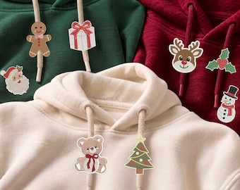 May include: Three hooded sweatshirts in dark green, deep red, and cream. Each hoodie features two drawstrings adorned with embroidered holiday-themed patches, including a gingerbread person, gift box, Santa, reindeer, holly, snowman, teddy bear, and tree.