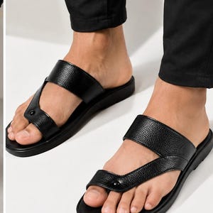 May include: A pair of black sandals featuring a textured leather-like finish, a wide strap across the midfoot, and a separate toe loop strap connected by a small circular stud detail. The sandals have a flat, solid black sole and an open-toe design.