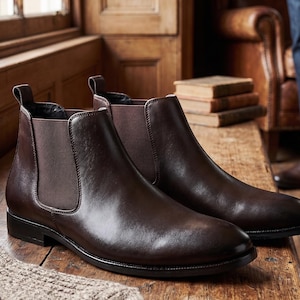 Op de afbeelding: Een paar donkerbruine leren Chelsea boots met een gladde afwerking, ronde neuzen, zwarte elastische zijpanelen en treklipjes aan de achterkant. De laarzen hebben een lage hak en een zwarte zool met een strak en eenvoudig ontwerp.