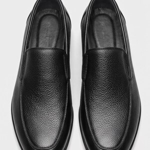 K&ouml;nnte beinhalten: Ein Paar schwarze Slipper mit einem strukturierten, genarbten Lederobermaterial. Diese Schuhe haben ein abgerundetes Zehendesign, ein glattes Innenfutter und eine klare, minimalistische Silhouette ohne Schn&uuml;rsenkel oder sichtbare Beschl&auml;ge.