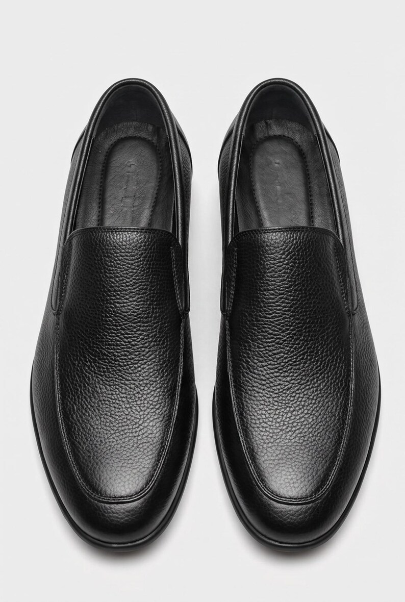 K&ouml;nnte beinhalten: Ein Paar schwarze Slipper mit einem strukturierten, genarbten Lederobermaterial. Diese Schuhe haben ein abgerundetes Zehendesign, ein glattes Innenfutter und eine klare, minimalistische Silhouette ohne Schn&uuml;rsenkel oder sichtbare Beschl&auml;ge.