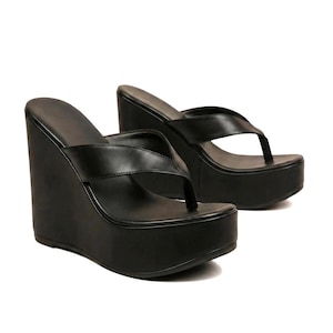 Peut inclure: Une paire de sandales compensées noires avec une bride entre-doigts. Ces chaussures présentent une semelle plateforme haute et une finition entièrement noire, avec une texture lisse sur les brides et la semelle intérieure, offrant un aspect uniforme.