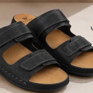 May include: A pair of black sandals with two adjustable hook-and-loop straps across the top. The footbed is tan, and the black straps feature visible stitching. The logo WALK CLASSIC CRAFTED FOR COMFORT is printed on the heel and the upper right.
