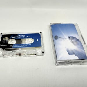 May include: A clear plastic cassette tape with a blue label featuring the text GEESE GETTING KILLED and the number 1. Beside it is a clear plastic cassette case with an insert showing a bright blue sky and a white bird in flight.