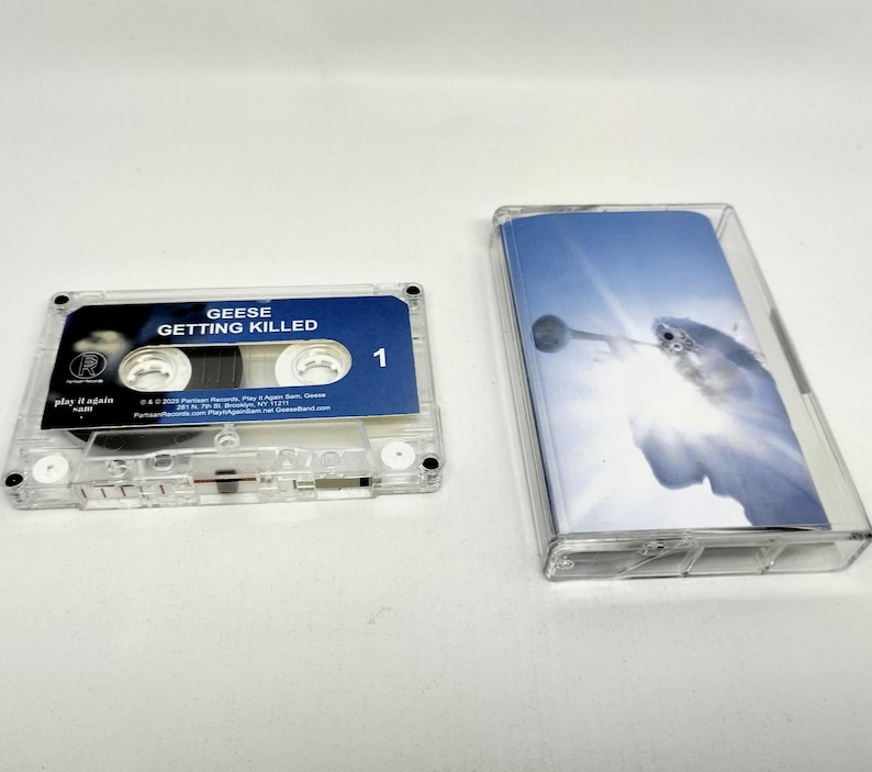 May include: A clear plastic cassette tape with a blue label featuring the text GEESE GETTING KILLED and the number 1. Beside it is a clear plastic cassette case with an insert showing a bright blue sky and a white bird in flight.