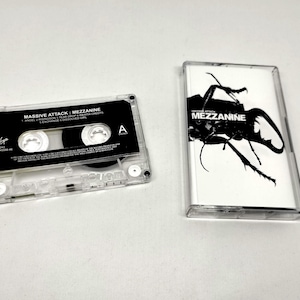 May include: A clear plastic cassette tape and its matching case for the album Mezzanine by Massive Attack. The cassette features a black label with white text, while the case insert displays a high-contrast black beetle on a white background.