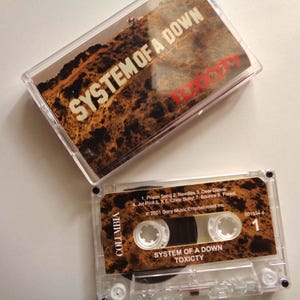 May include: A clear cassette tape and case featuring the album "Toxicity" by System of a Down. The cassette has a brown and tan design with the band's name and album title. The case also displays the album title.