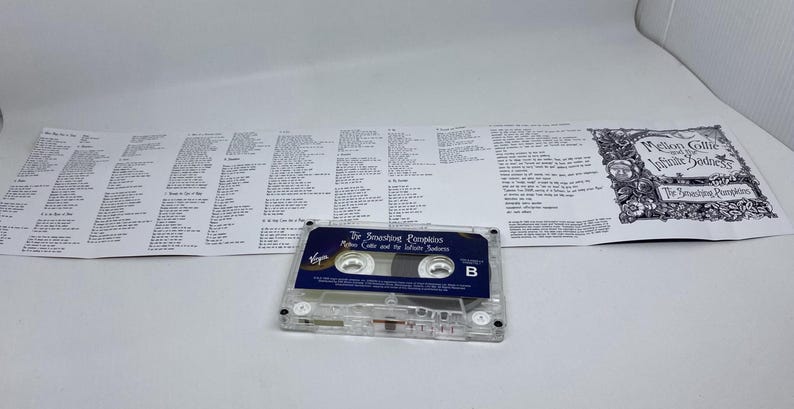 Pu&ograve; includere: Una musicassetta trasparente con un'etichetta blu e bianca, con il testo "The Smashing Pumpkins Mellon Collie and the Infinite Sadness". La musicassetta &egrave; accanto a un foglio di testi piegato con titoli di canzoni e illustrazioni.