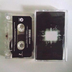 May include: A clear cassette tape case with a black cassette inside. The cassette has white text that reads "TOOL ÆNIMA" and track titles. The case is next to a black cassette cover with a white design.