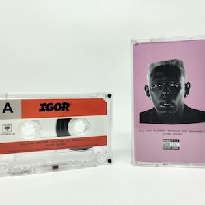 Puede incluir: Un casete transparente con la palabra "IGOR" en una etiqueta roja, junto a una caja de casete con un fondo rosa y un retrato en blanco y negro. El texto "ALL SONG WRITTEN, PRODUCED AND ARRANGED BY TYLER OKONMA" es visible.