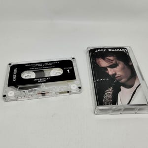 May include: A clear cassette tape labeled "Jeff Buckley Grace" next to its case, featuring a black and white image of a man. The cassette tape has a black label with white text and the Columbia logo.