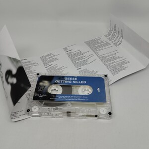 May include: A clear plastic cassette tape with a blue label featuring the text Geese Getting Killed and the number 1. An unfolded white paper j-card insert with black printed lyrics and credits is positioned behind the cassette tape.