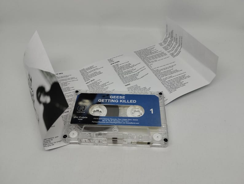 May include: A clear plastic cassette tape with a blue label featuring the text Geese Getting Killed and the number 1. An unfolded white paper j-card insert with black printed lyrics and credits is positioned behind the cassette tape.