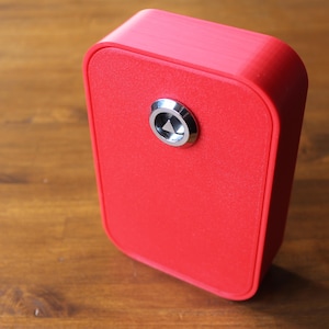 May include: A bright red, rectangular box with rounded corners and a silver button. The box is positioned at an angle on a wooden surface, showcasing its design and color. The button is centered on the front.