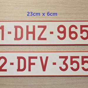 May include: Two rectangular white license plates with red borders and red embossed characters. The top plate reads 1-DHZ-965 and the bottom plate reads 2-DFV-355. Each plate features four small corner holes and measures 23 cm by 6 cm in size.