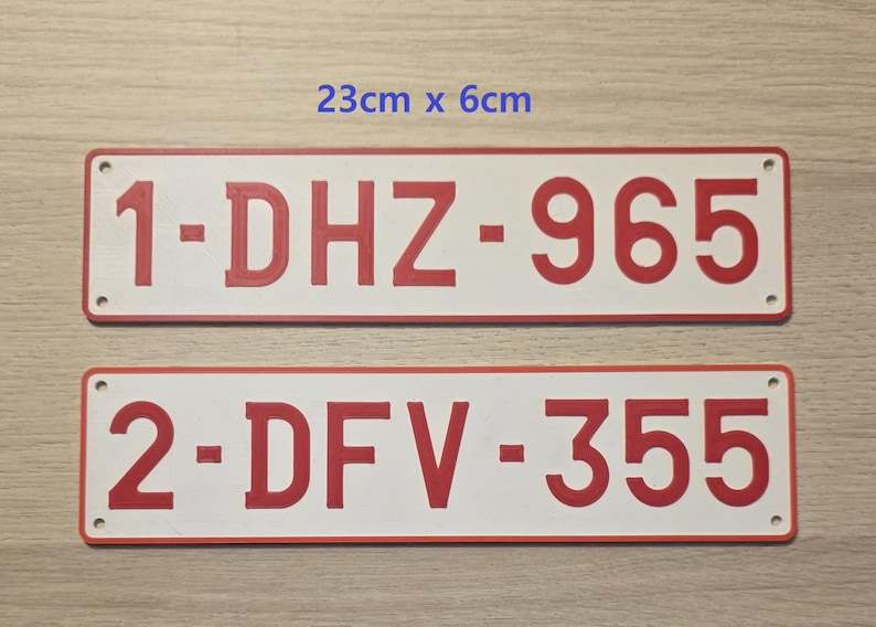 May include: Two rectangular white license plates with red borders and red embossed characters. The top plate reads 1-DHZ-965 and the bottom plate reads 2-DFV-355. Each plate features four small corner holes and measures 23 cm by 6 cm in size.