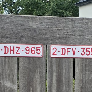 May include: Two rectangular white metal license plates with red borders and red lettering. The left plate displays the alphanumeric code 1-DHZ-965, and the right plate displays the alphanumeric code 2-DFV-355. Both plates feature small mounting holes.