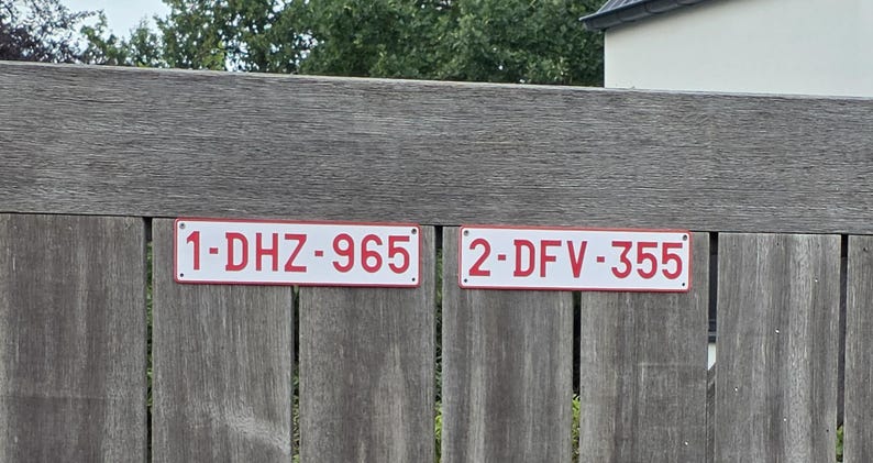 May include: Two rectangular white metal license plates with red borders and red lettering. The left plate displays the alphanumeric code 1-DHZ-965, and the right plate displays the alphanumeric code 2-DFV-355. Both plates feature small mounting holes.