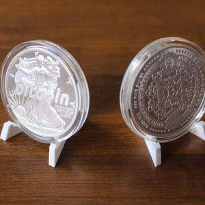 May include: Two silver coins in clear protective cases, displayed on small white stands. One coin features a woman with the word "Bitcoin" and the other has an intricate design. The coins are set against a dark wood surface.
