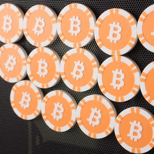 May include: A collection of orange and white Bitcoin-themed tokens. Each token features a white "B" symbol in the center, surrounded by a white and orange striped border. The tokens are arranged on a dark, textured surface.