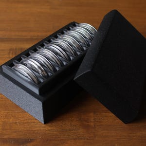 May include: A black box with a foam insert holds multiple silver coins. The box is open, revealing the coins neatly arranged. The lid of the box is slightly ajar, and the background is a wooden surface.