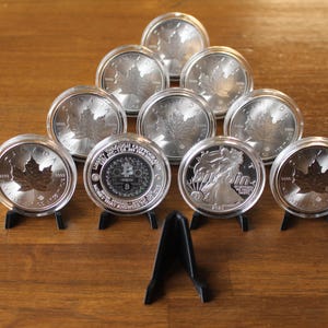 May include: A collection of silver coins, some featuring a maple leaf design, displayed on small black stands. The coins are arranged in a pyramid shape on a wooden surface, showcasing their intricate details and reflective surfaces.
