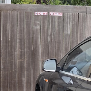 May include: A vertical wooden fence featuring two rectangular white signs with red borders and text. The left sign displays 1-DHZ-965 and the right sign displays 2-DFV-355. A portion of a black car is visible in the foreground, showing the side mirror.