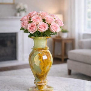 May include: A decorative vase filled with pink roses. The vase is made of green and gold-toned onyx, with a flared top and a pedestal base. The roses are in full bloom, creating a vibrant floral arrangement. The vase is placed on a marble-topped table.