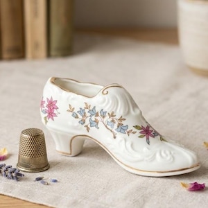 May include: A decorative white porcelain shoe with pink and blue floral patterns and gold detailing. A vintage metal thimble is placed next to the shoe. Dried lavender and rose petals are scattered nearby.