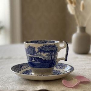 May include: A blue and white ceramic teacup and saucer set with a landscape design. The cup has a unique handle and sits on a matching saucer. The set is placed on a white tablecloth.
