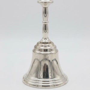 Puede incluir: Una campana plateada con una parte superior redondeada y un mango largo. La campana tiene una base acampanada con crestas horizontales e hendiduras verticales. El mango tiene una parte superior bulbosa y un diseño delgado y acanalado.